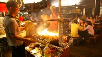 Exterior night shot of men barbequing satay with people dining